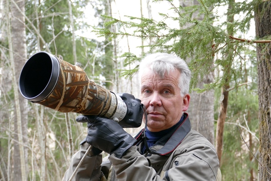 Lähikuva miehestä, joka pitelee kameraa. Kamerassa on pitkä teleobjektiivi. Taustalla näkyy metsä.