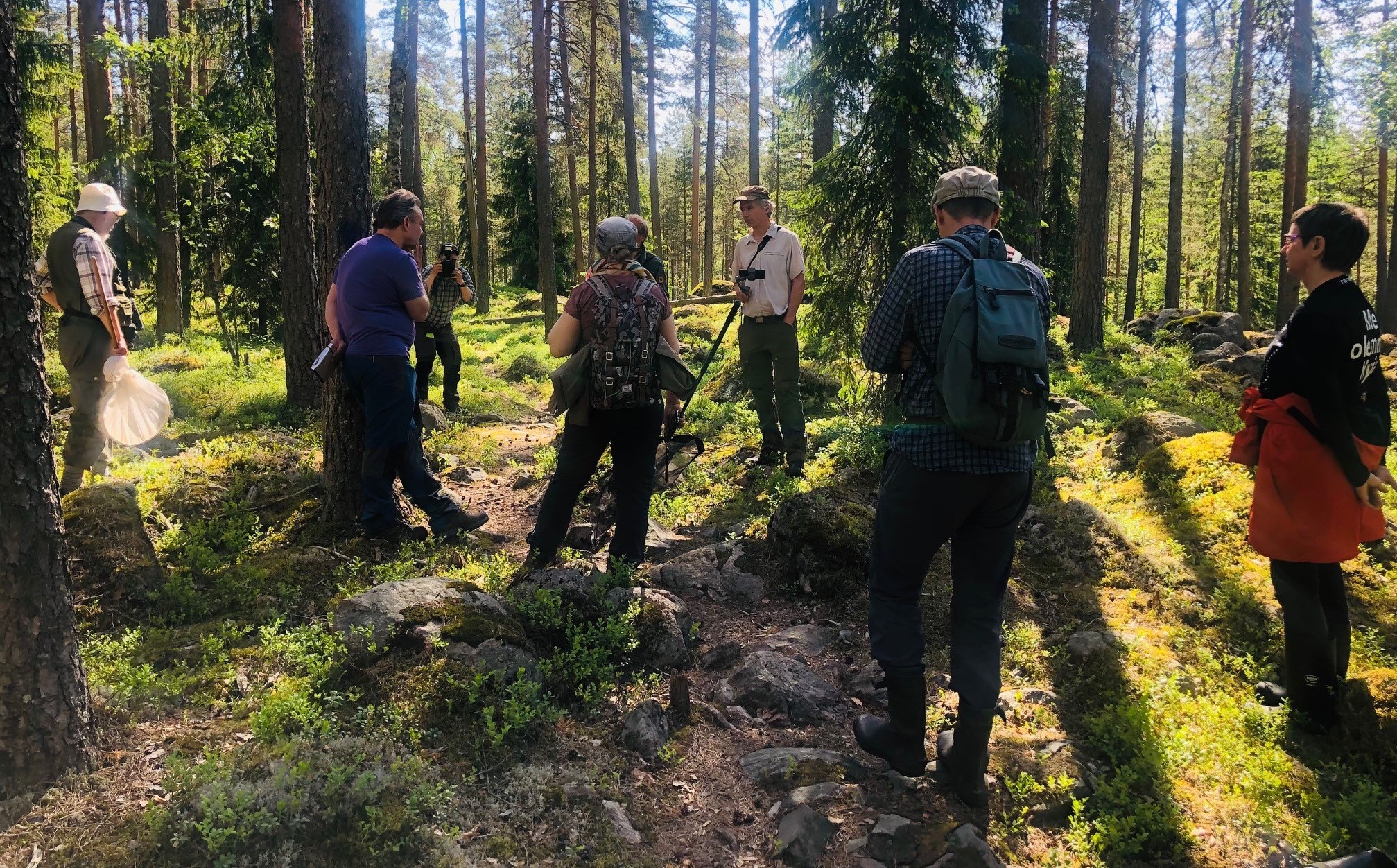 Joukko ihmisiä seisoo harvapuutoisessa metsässä.