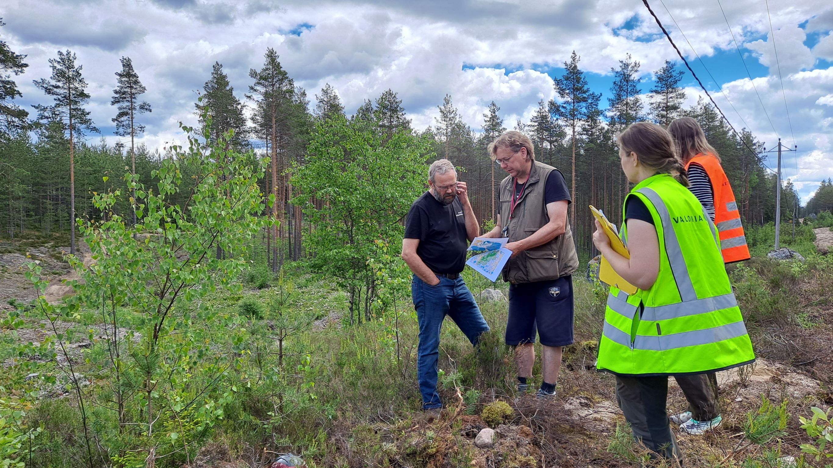 Neljä ihmistä kartan tutustumassa karttaan. Ympärillä nuorta lehtipuustoa ja taustalla vanhempaa männikköä. Sininen taivas pilvipoutainen.